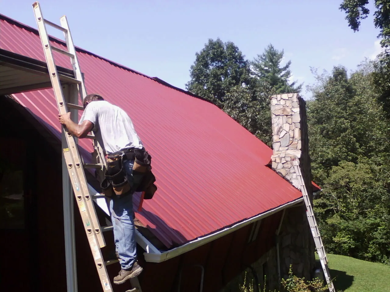 Roofer on a ladder beside a red metal roof and stone chimney — Soffit & Fascia Repair by Roofing Brothers in Bell Buckle